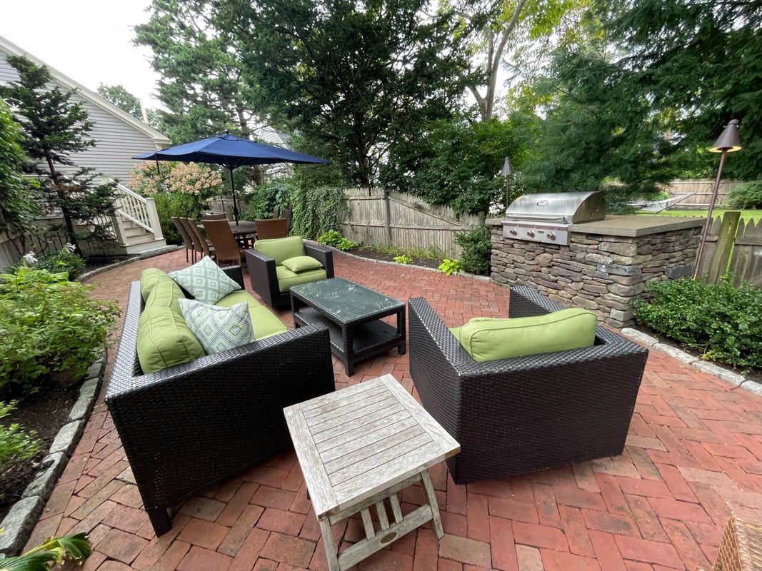 Patio with wicker furniture, a grill, and a blue umbrella. Brick patio with greenery surrounds.