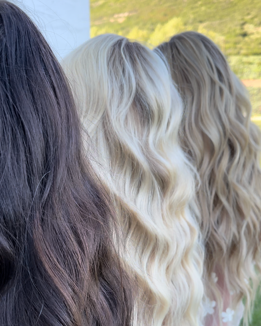 Three women with long, wavy hair in shades of dark brown, blonde, and light brown. They are outside in a natural setting.