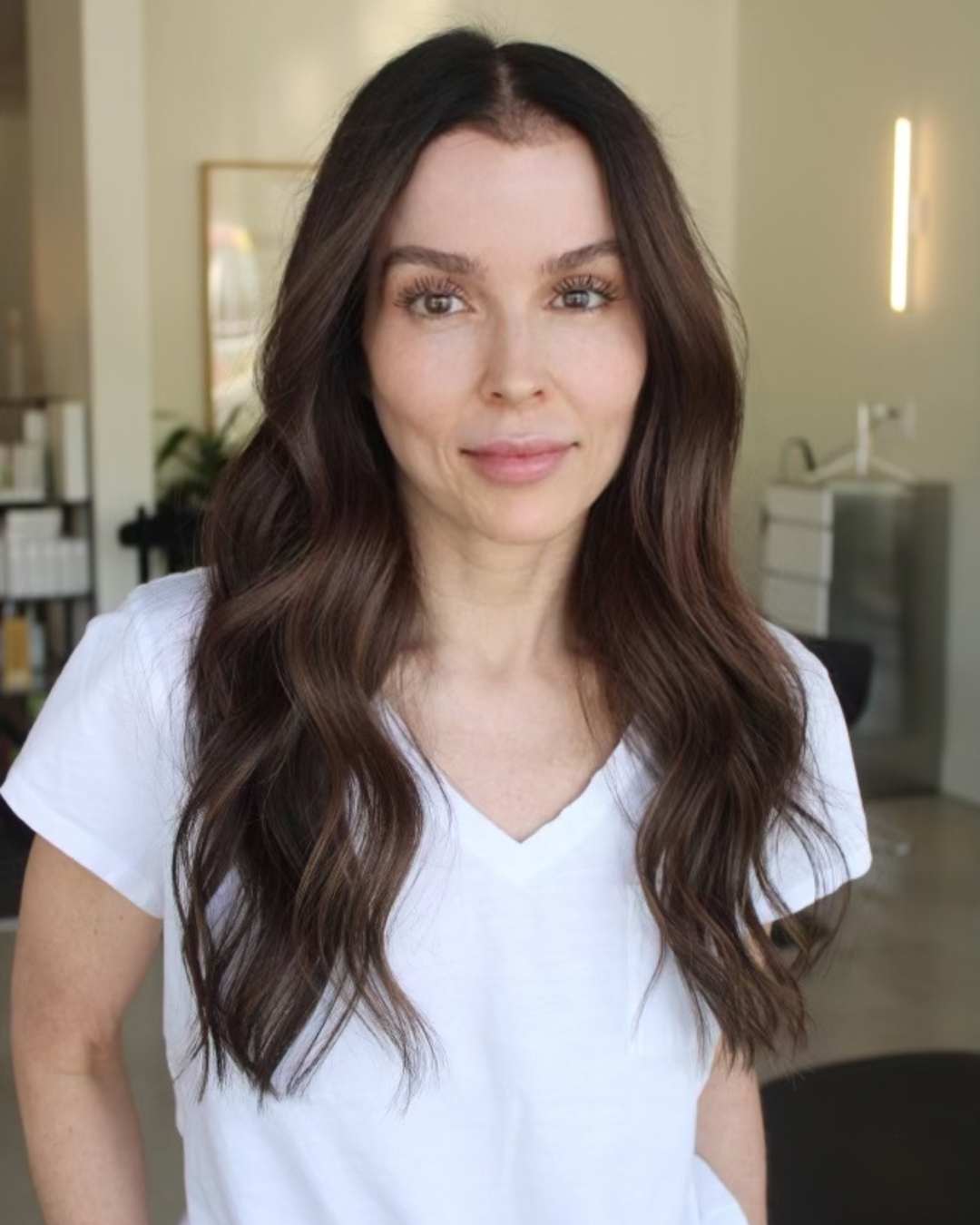 Woman with long, wavy brown hair and a white v-neck shirt smiles at the camera indoors.