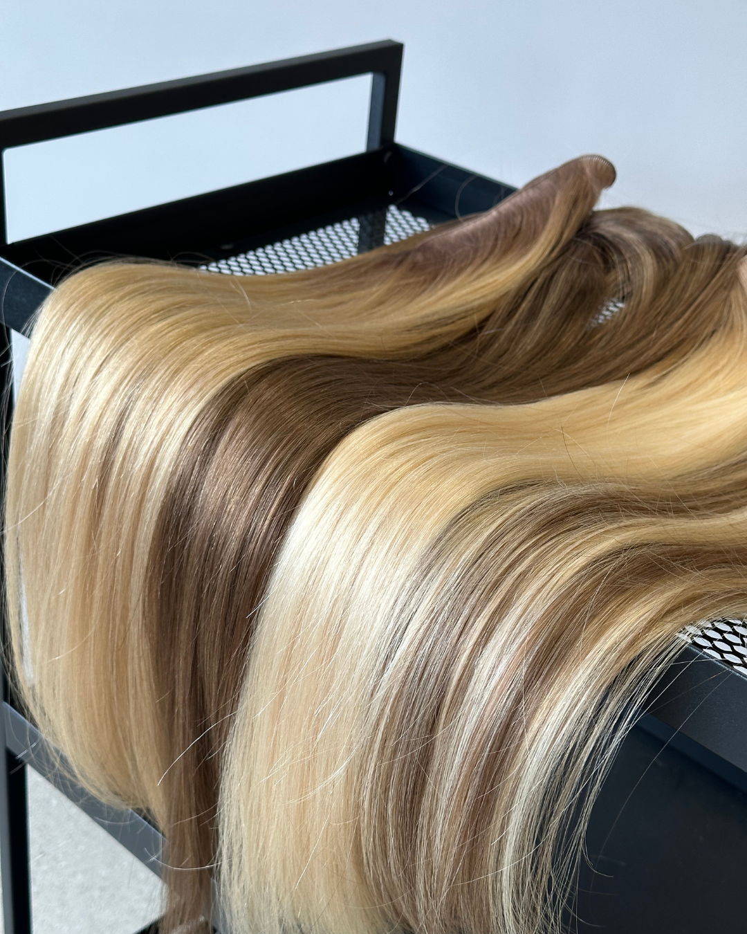 Hair extensions in varying shades of blonde and brown, displayed on a black rack, with highlights visible in the light.
