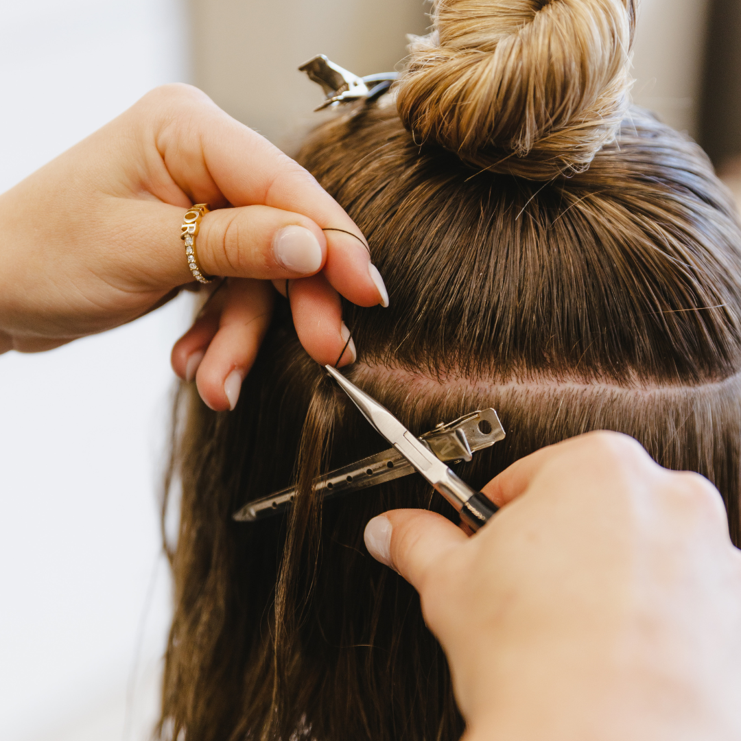 Hands installing hair extensions on a person. One hand holds a small tool, while the other parts hair.
