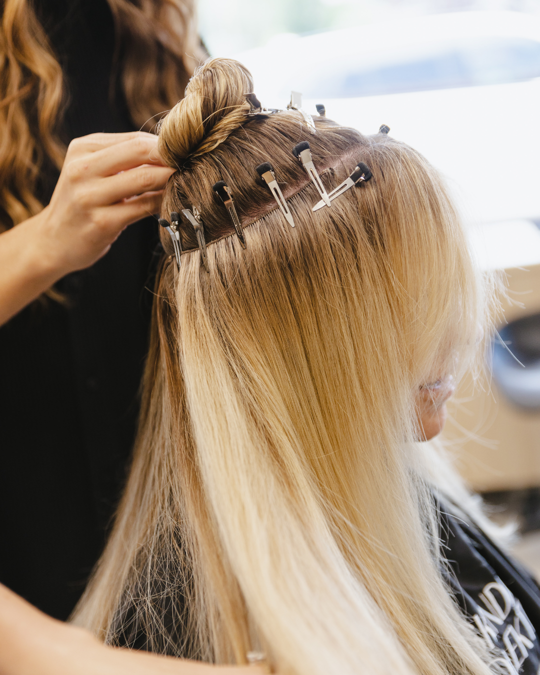 A hairstylist installing blonde hair extensions on a person with blonde hair in a salon.