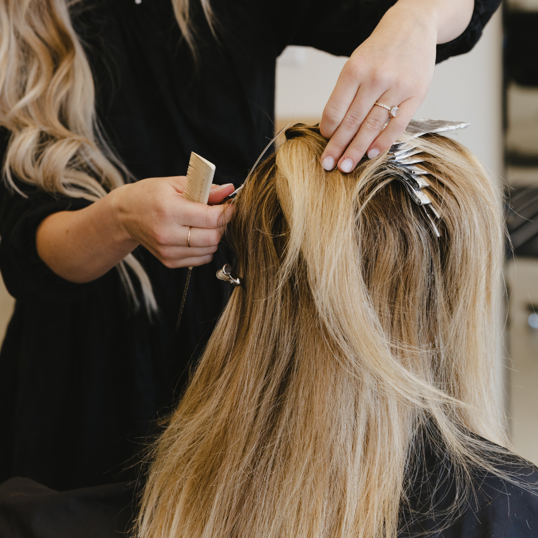 A hairstylist is applying highlights to a client's long blonde hair in a salon.