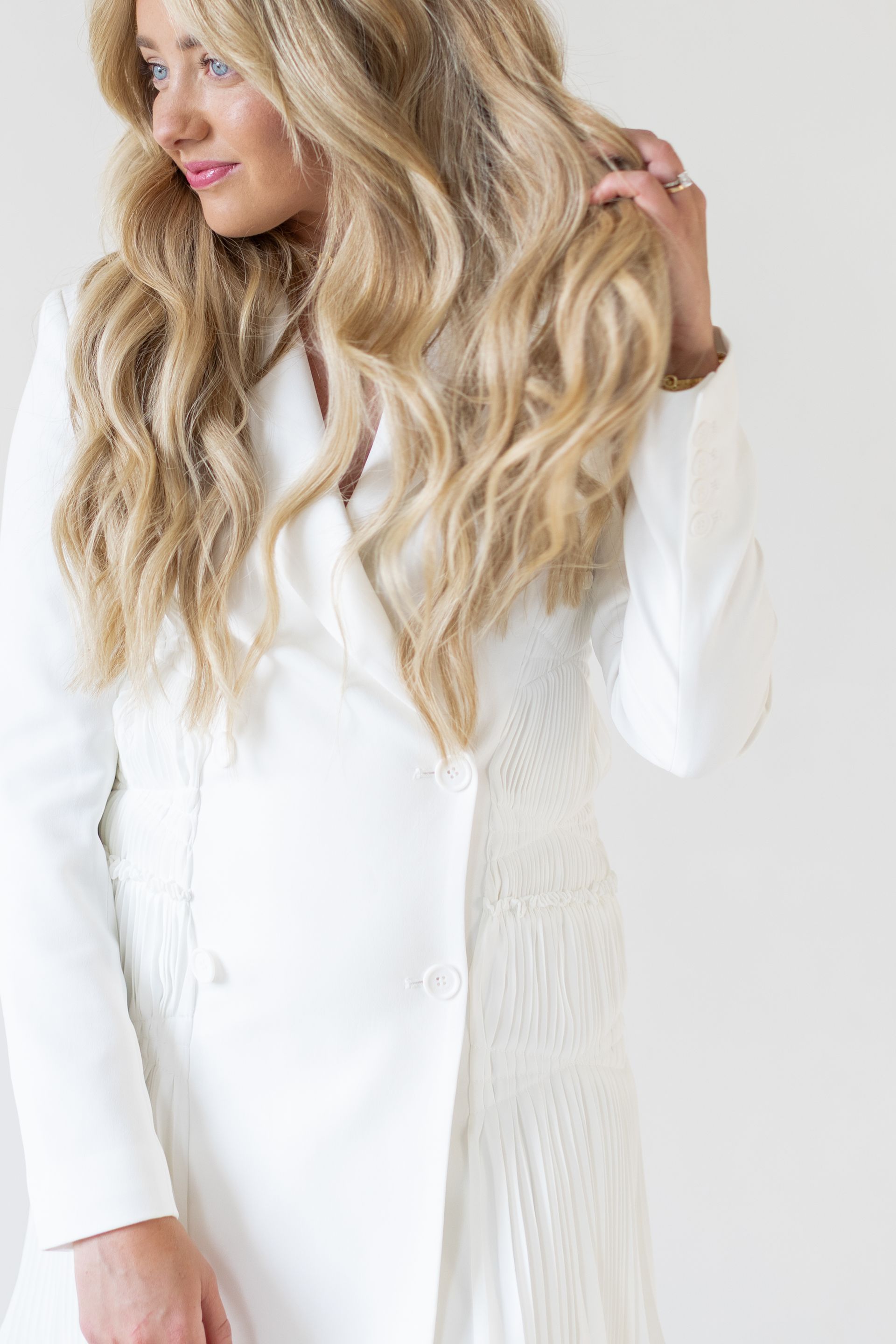 Blonde woman in white blazer, hand in hair, smiling against a white backdrop.