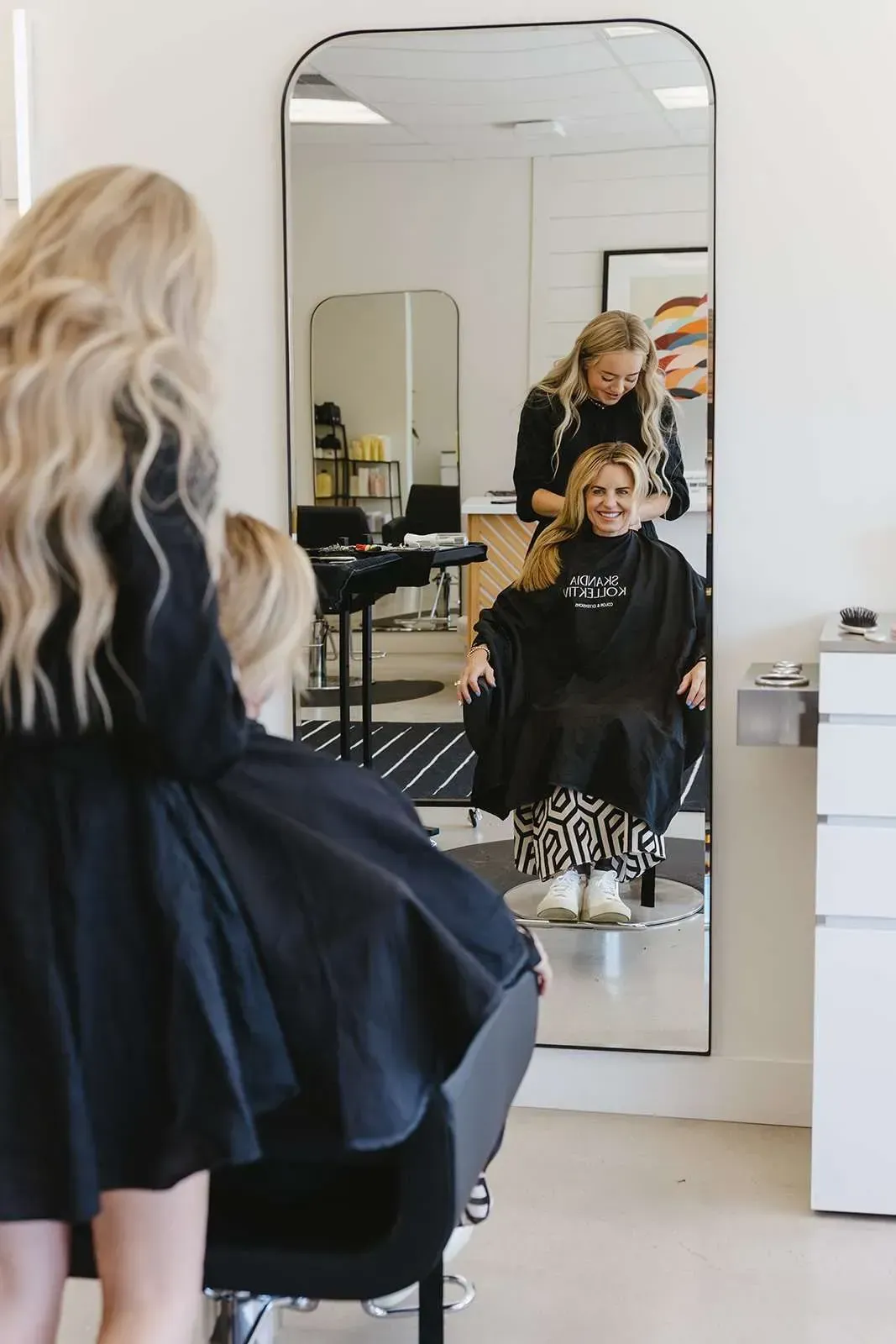 Two blonde stylists work on a young girl's hair in a salon, one facing the girl, the other reflected in a tall mirror.