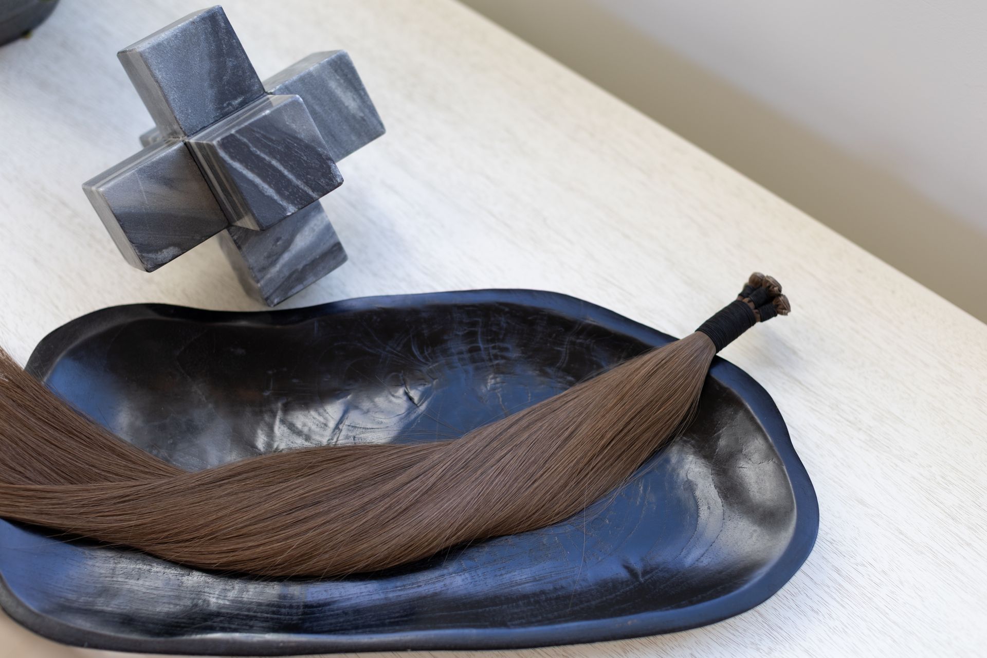 Brown hair extensions in a black, oval bowl; gray decorative piece on a light surface.