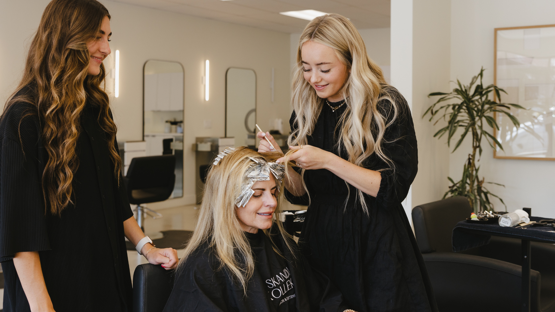 Three women at a hair salon. A stylist foils a client's light blonde hair, while another woman looks on and smiles.