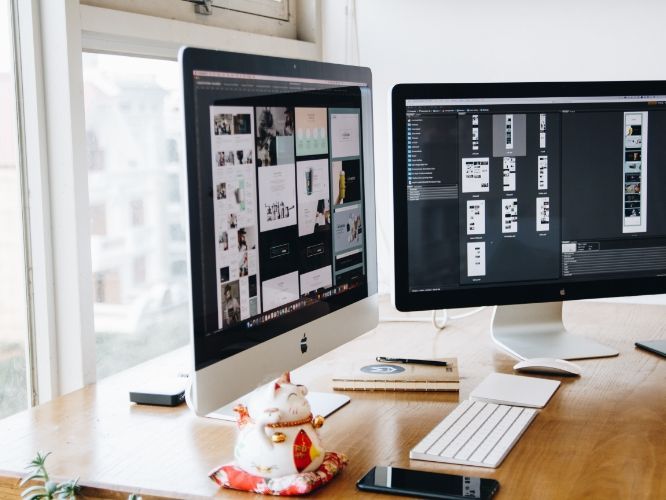 Two computer monitors displaying design work on a wooden desk near a window. A lucky cat figurine sits in the foreground.