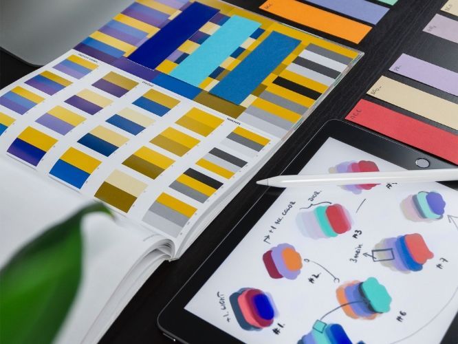 Color swatches and sketches on a tablet and in a notebook, indicating design choices. A stylus rests on the tablet.