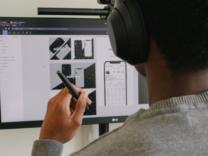 A person with headphones designs on a computer, using a stylus to interact with mockups of a mobile app interface.A