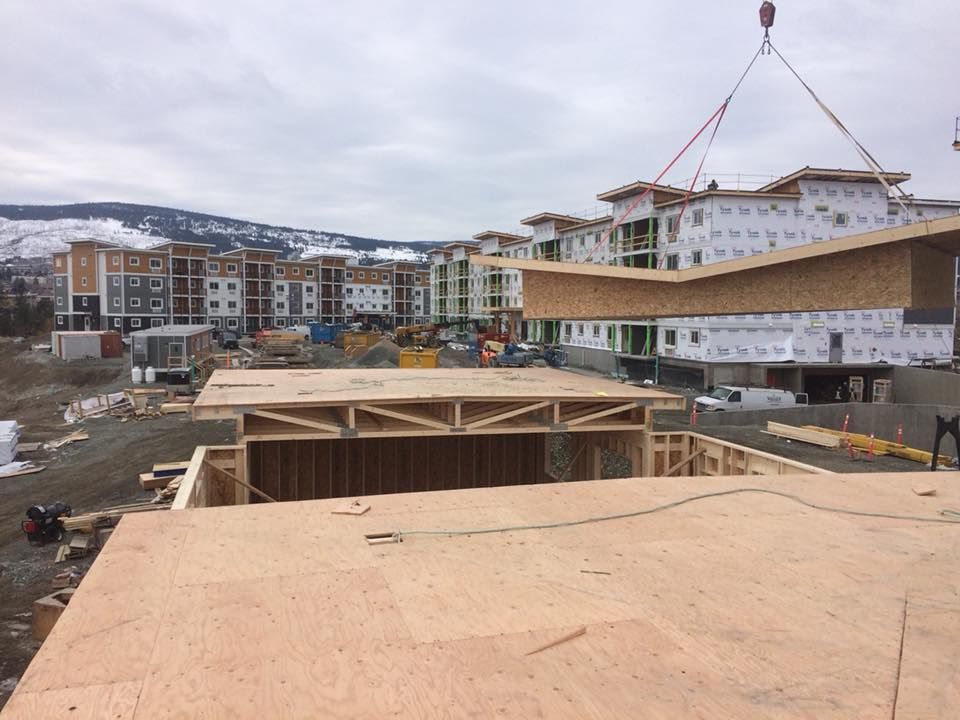 crane lifting a roof section onto a partially built apartment building, with other buildings