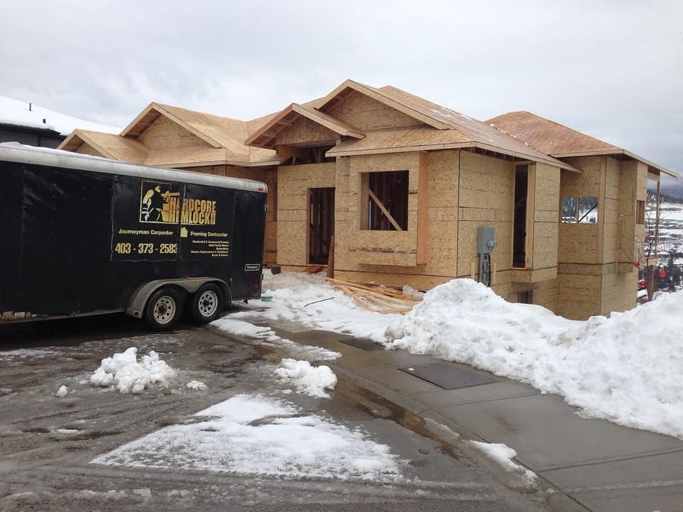 Construction of a wood-framed house with snow on the ground and a black trailer.