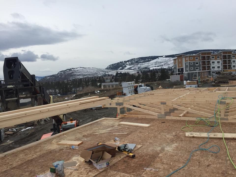 Construction site with crane lifting a wooden beam onto a roof frame; mountains in the background.