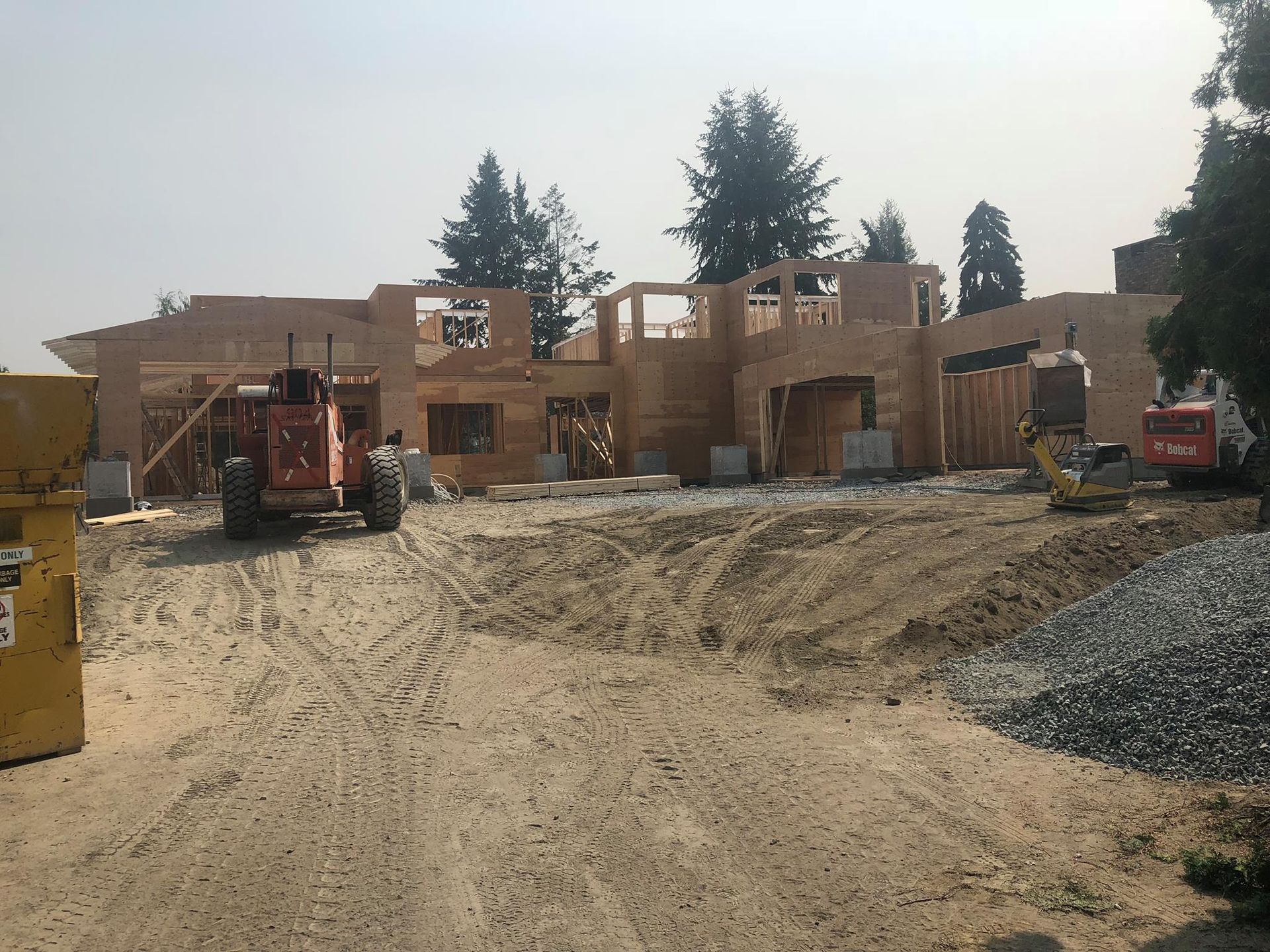Construction site: wood-framed house under construction with heavy machinery on dirt. Trees in the background.