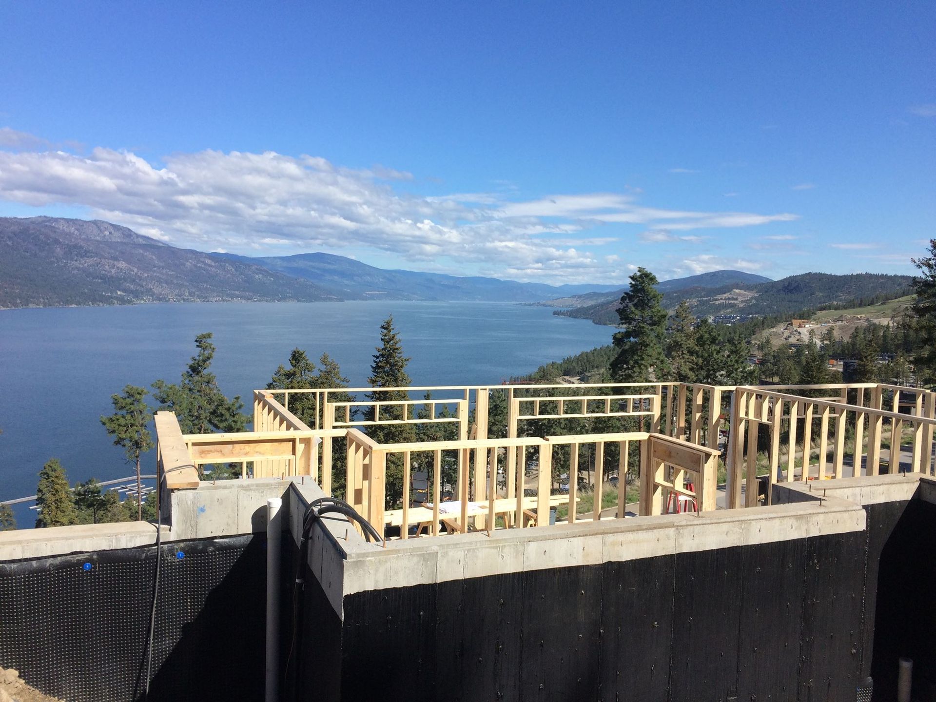 Construction of a house with wooden frames overlooking a lake and mountains under a blue sky.