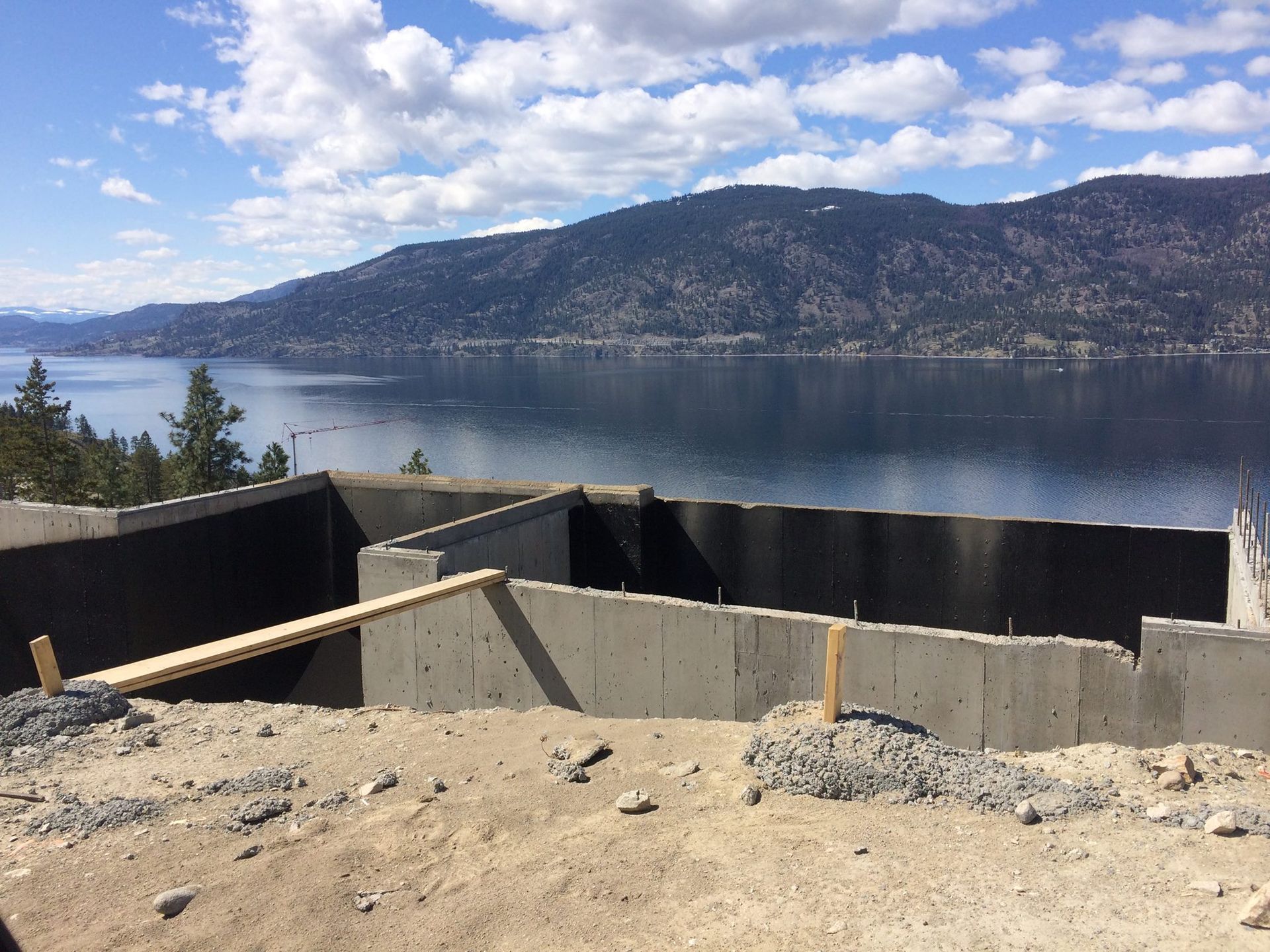 Foundation walls under construction, overlooking a lake and mountains on a sunny day.