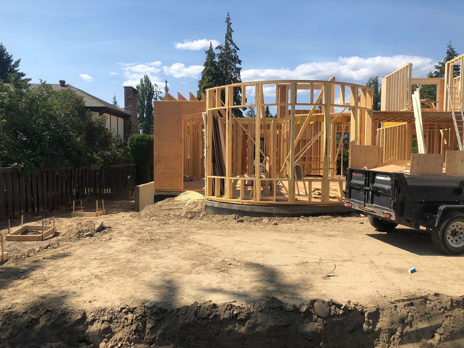 Wooden house frame under construction outdoors on a sunny day; a trailer is parked nearby.