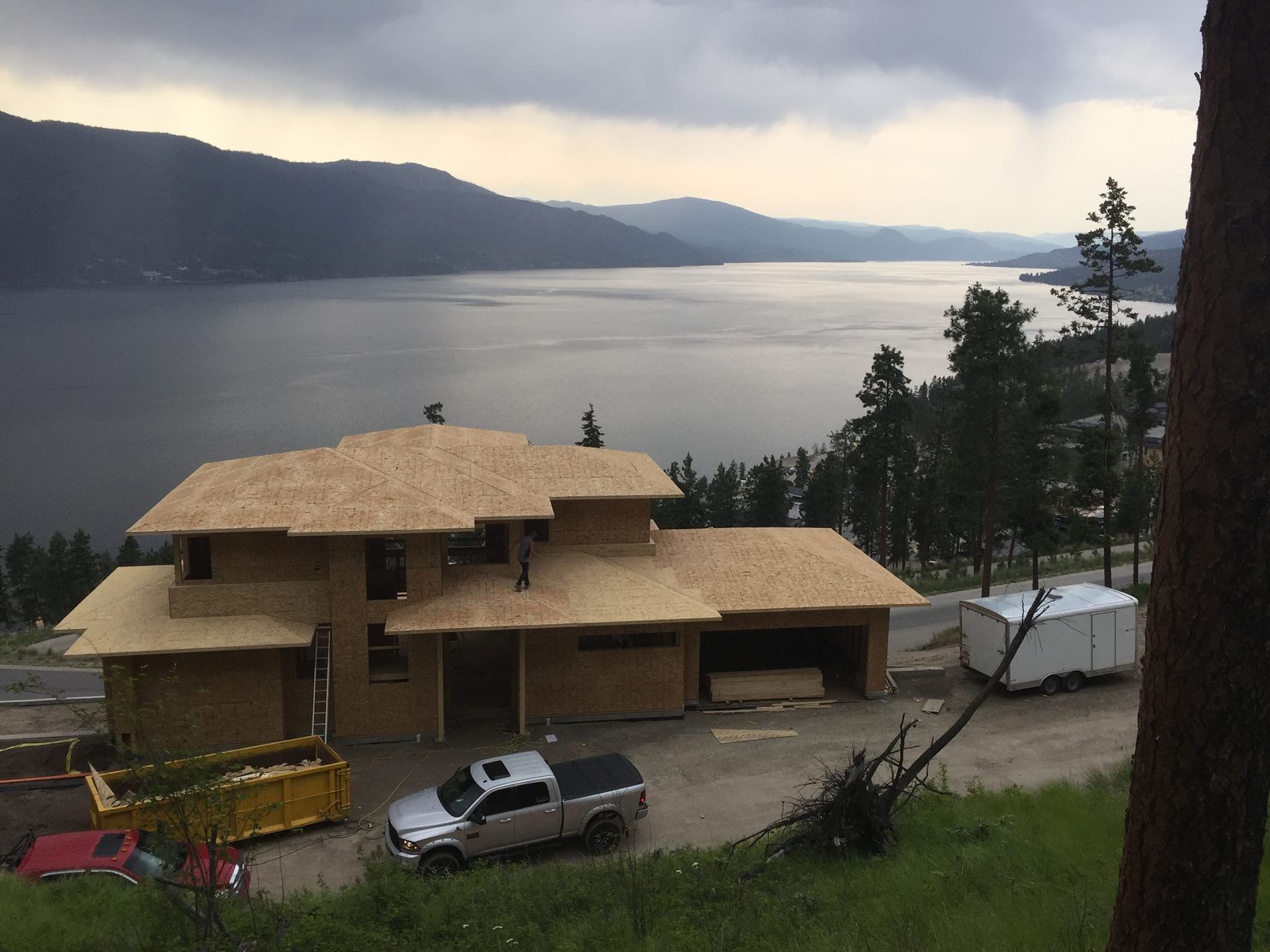 House under construction overlooking a lake and mountains; workers on roof, cloudy sky.