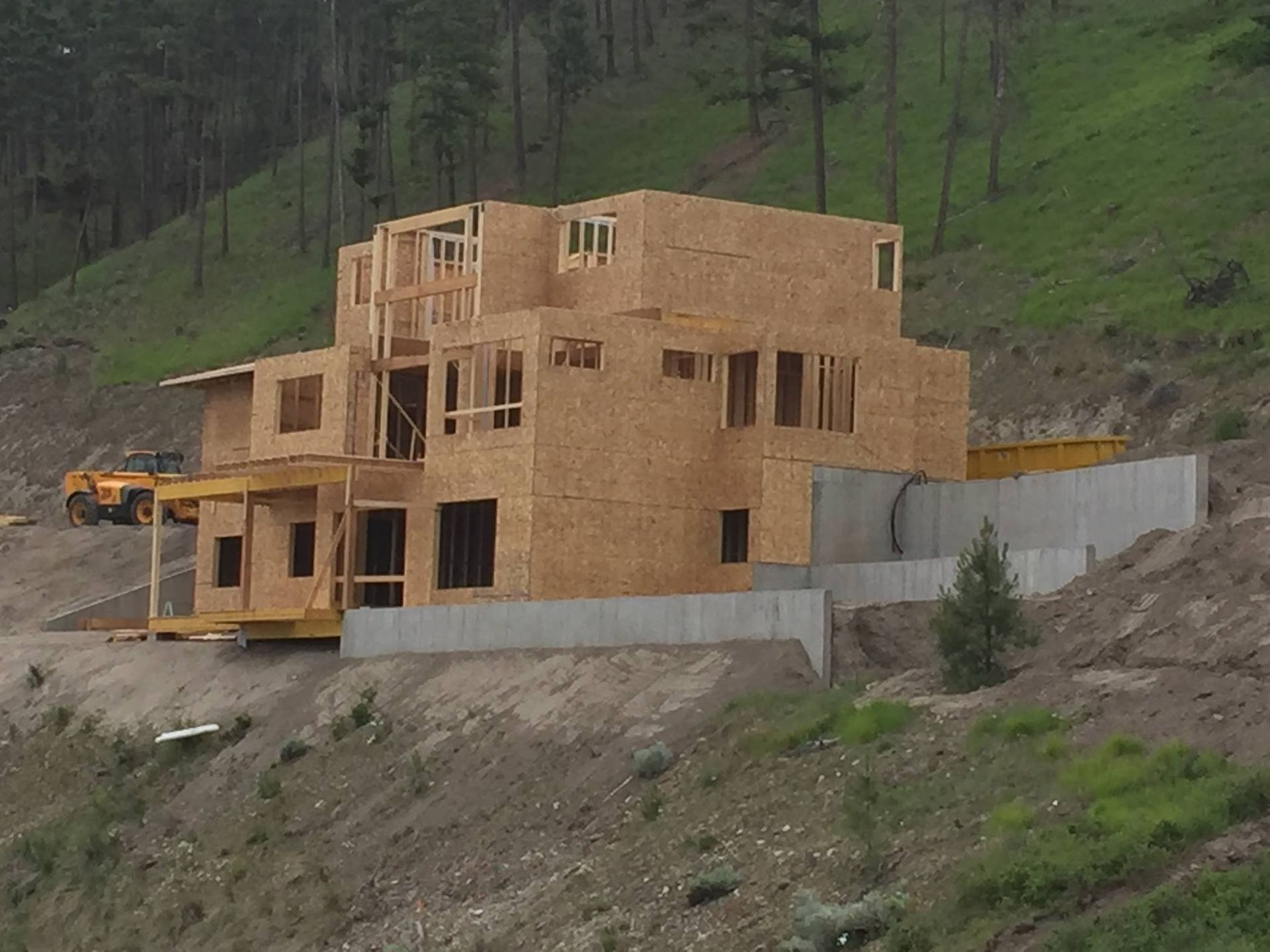 House under construction on a hillside, framed with plywood. Concrete retaining wall.