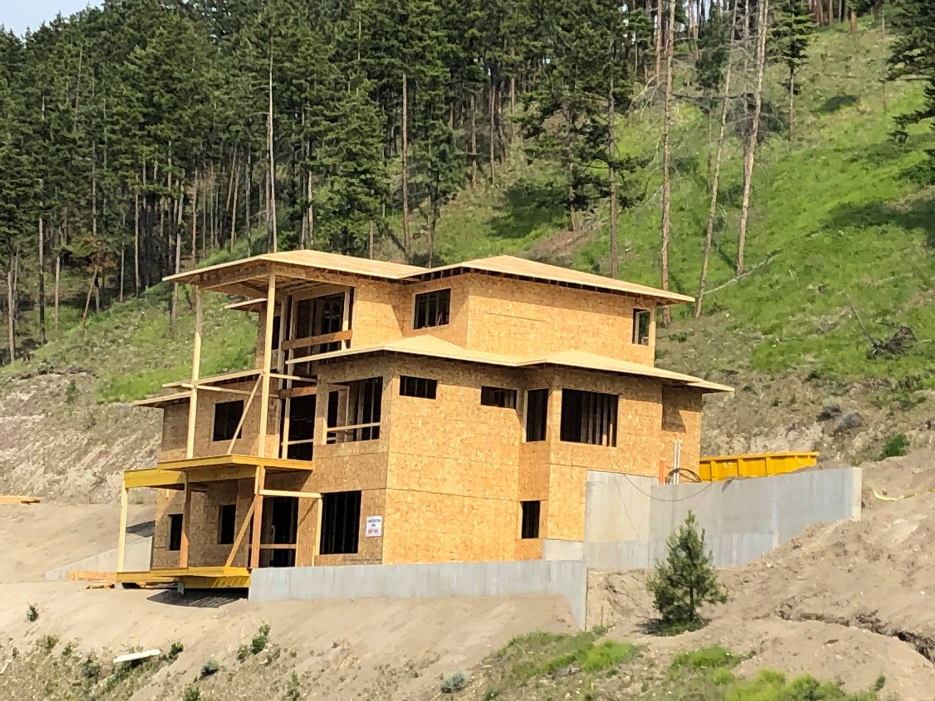 House under construction, two stories, on a hillside, surrounded by forest and retaining walls.