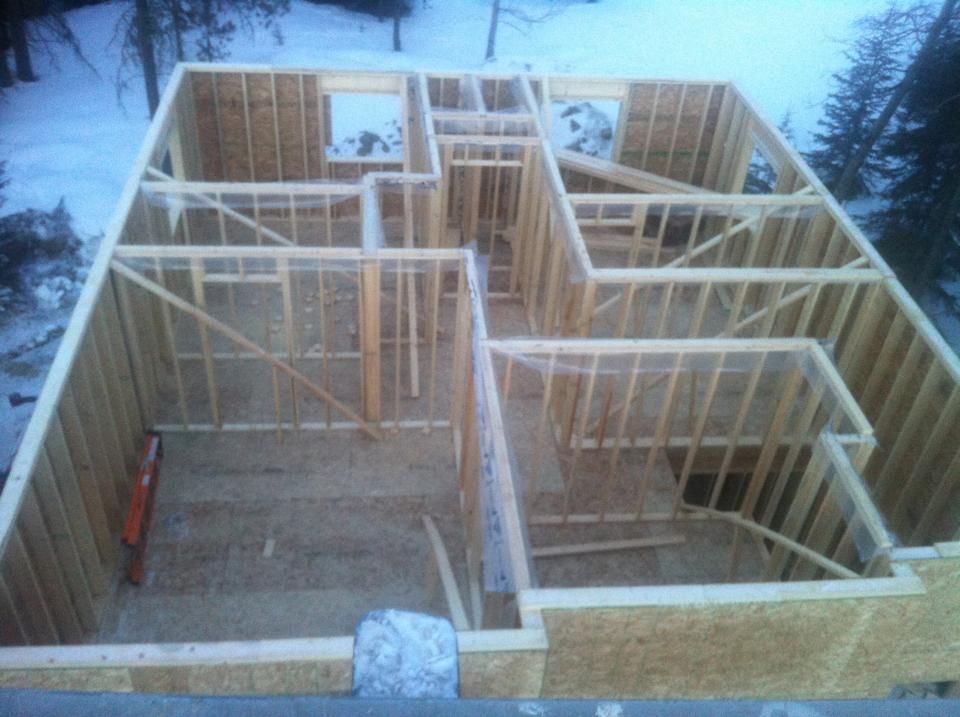 Wooden frame of a house under construction; walls, windows, and a staircase are visible, with a snowy background.