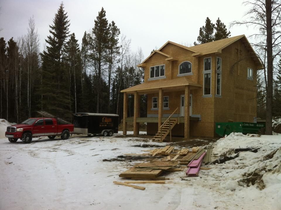 A two-story house under construction in a snowy, wooded area; a red truck and trailer are parked nearby.