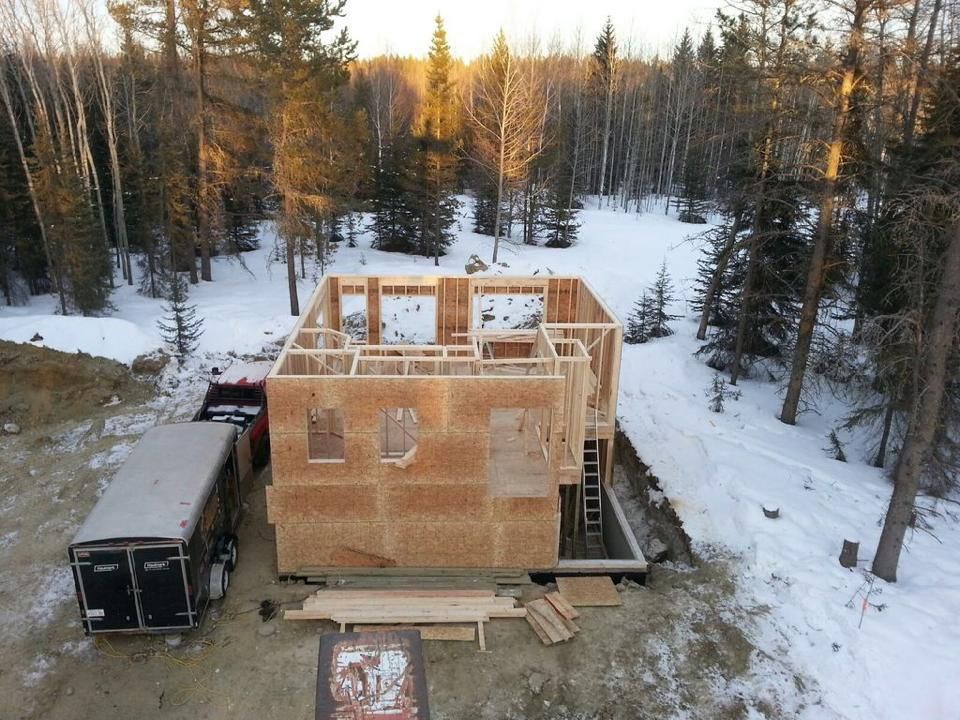 Construction of a wooden house in a snowy, forested area. A trailer and truck are parked nearby.