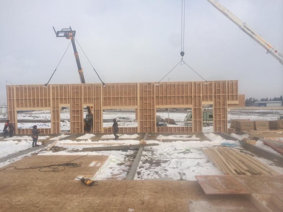 Construction workers using cranes to lift wooden wall panels onto a snowy building site.