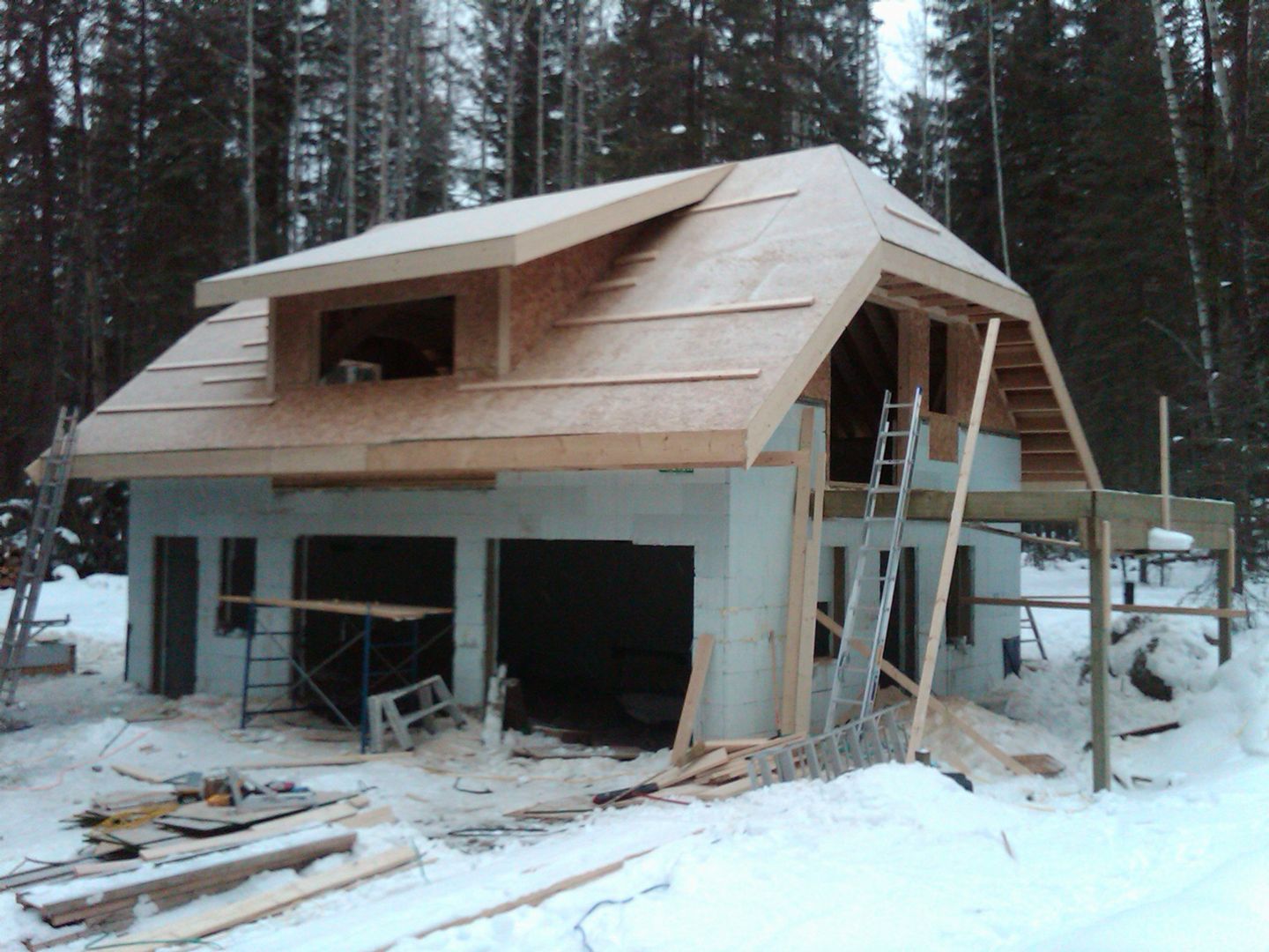 Construction of a two-story building in a snowy forest. The roof and dormer are wood, the walls are light gray.