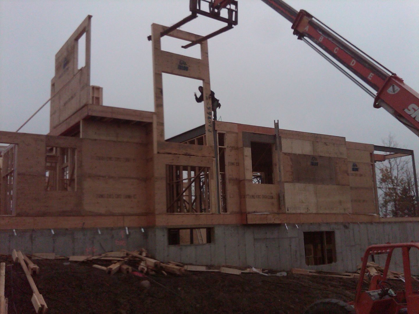 Construction site: crane lifting wooden wall panel onto concrete foundation. Cloudy sky.