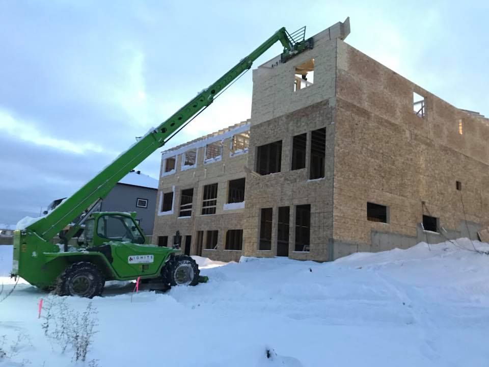 Green telehandler extends to top of a multi-story building under construction on snow-covered ground.