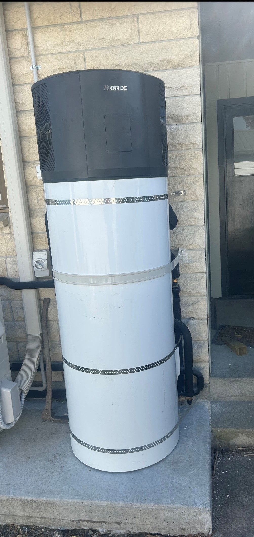 Large, cylindrical, white water heater with black top, outdoors on a concrete slab, near a brick wall.