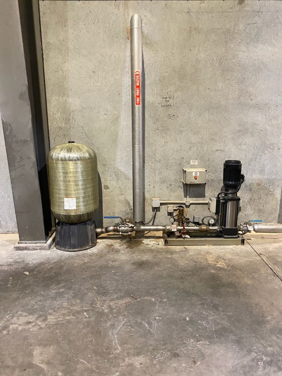 Industrial equipment against a concrete wall: a pressure tank, a pump, and a tall, labeled pipe.