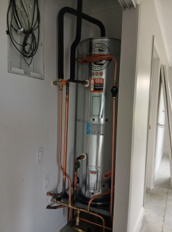 A tall silver water heater with copper pipes in a white closet. Black pipes connect to the top.