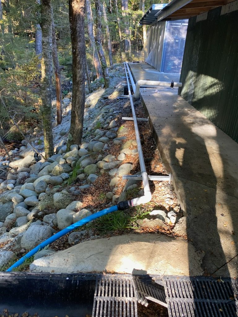 A concrete pathway with a drain and water hose, next to a rocky slope and trees.