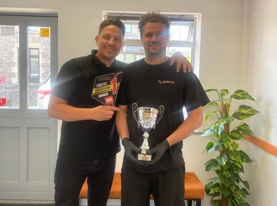 Two smiling individuals stand side-by-side in a room, with one holding a silver trophy while wearing black gloves.