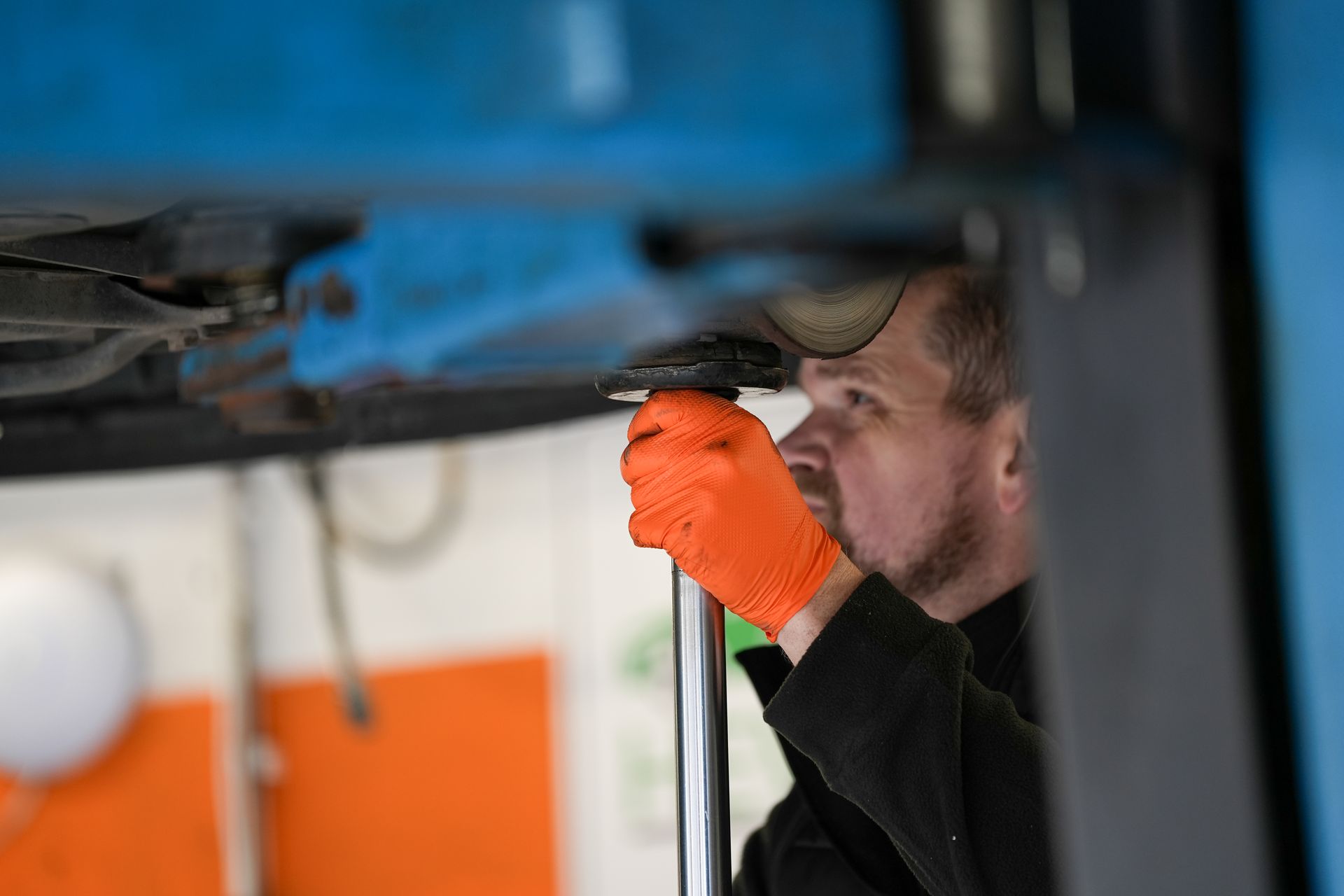 Two technicians in matching blue uniforms examine the open engine bay of a metallic orange car in a bright auto shop.