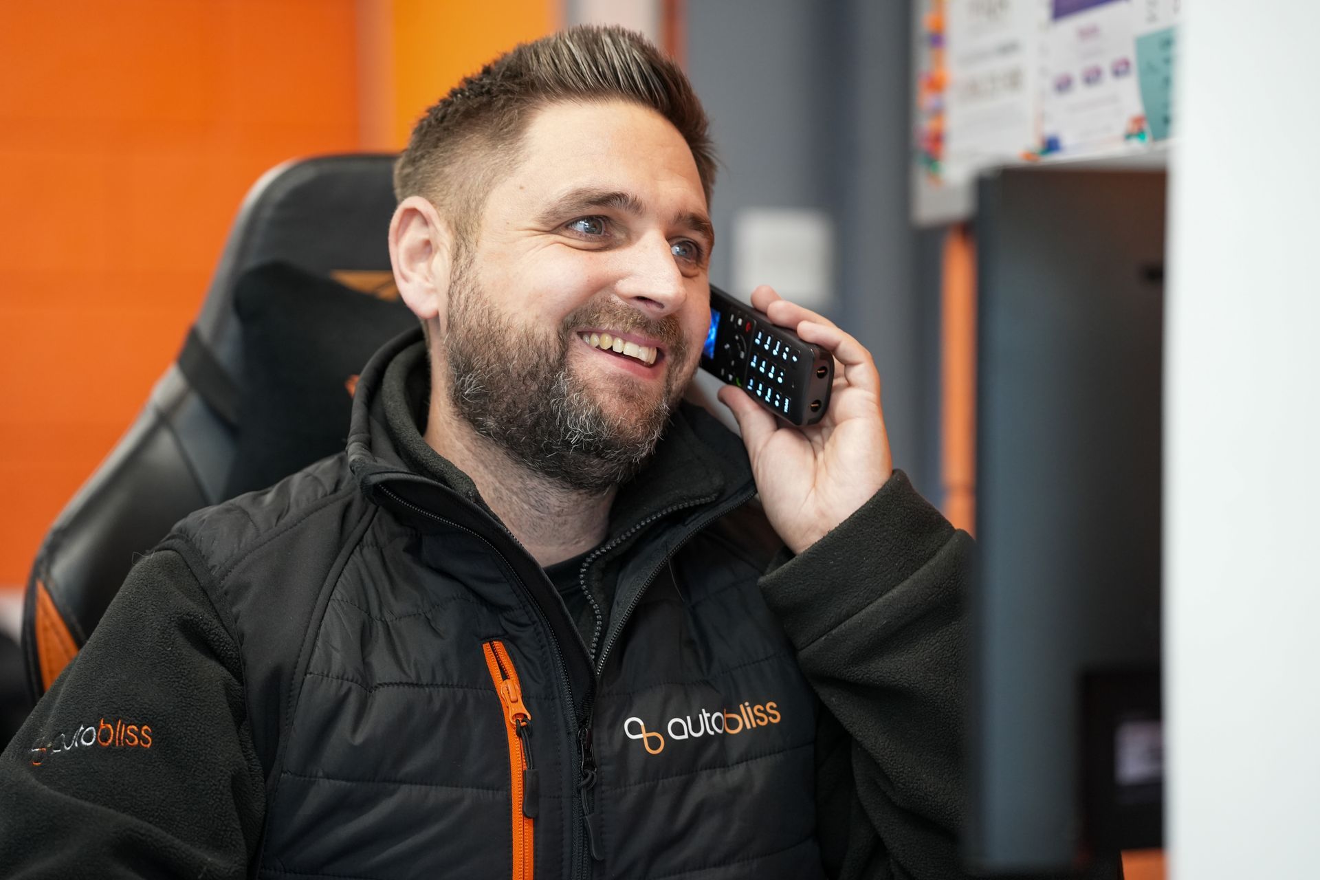 Two technicians in matching blue uniforms examine the open engine bay of a metallic orange car in a bright auto shop.