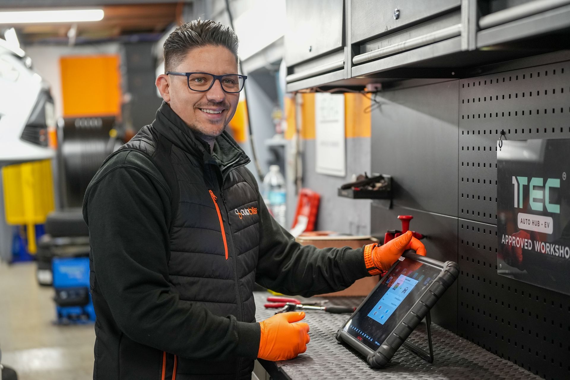 A smiling mechanic in a workshop examines a car engine with a diagnostic tool, wearing a black vest and orange gloves.