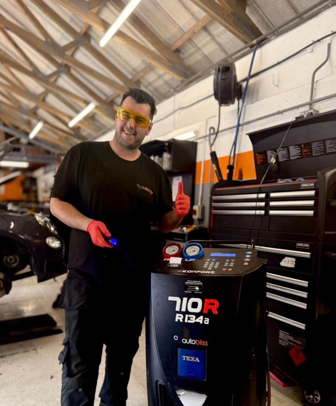 Hands in bright orange gloves hold an automotive diagnostic scan tool with glowing indicator lights on a dark surface.