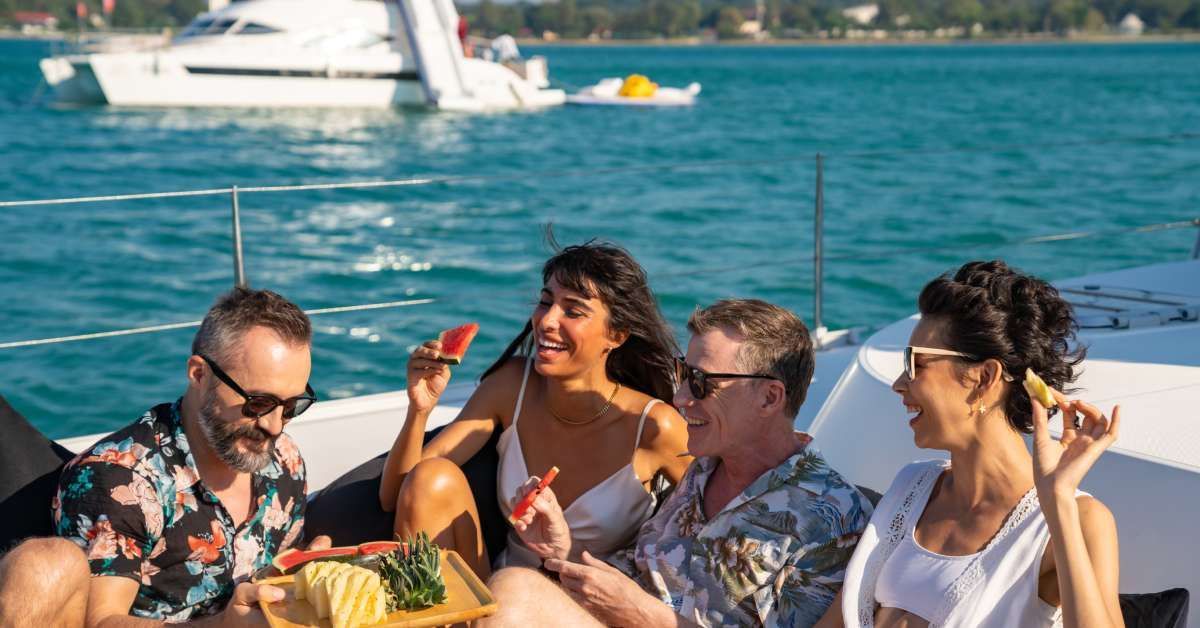 Group of men and women friends enjoying an outdoor party, eating fresh fruit together while a catamaran boat is sailing.