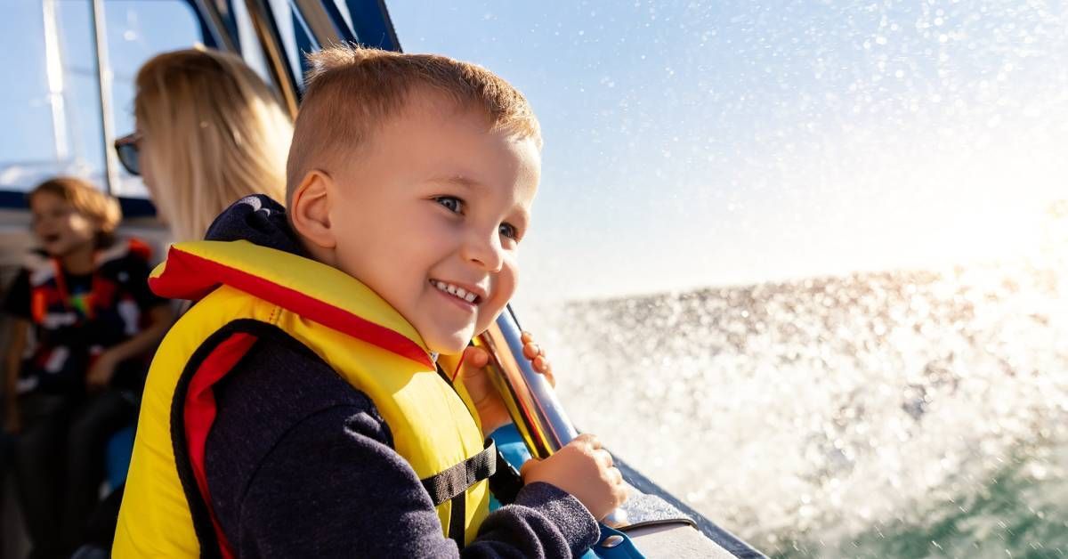 Happy little blond boy wearing a life vest, smiling with excitement as he enjoys sailing on a motorboat.