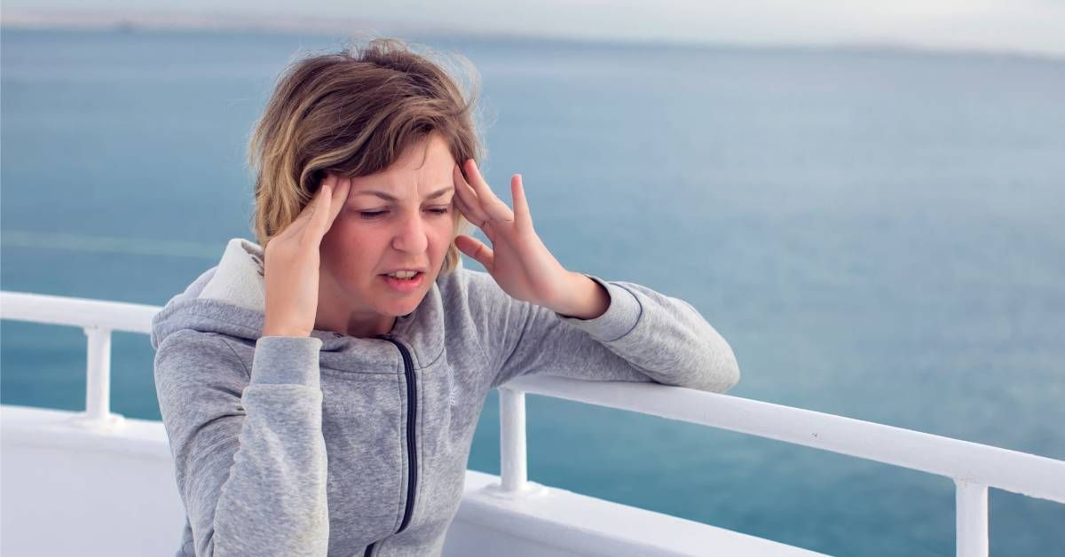 Woman in a gray jacket suffering from seasickness on a cruise ship, holding her head and covering her mouth.