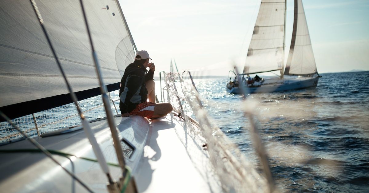 The backside of a man sitting on a sailboat. The man watches another sailboat move across the water in the distance.