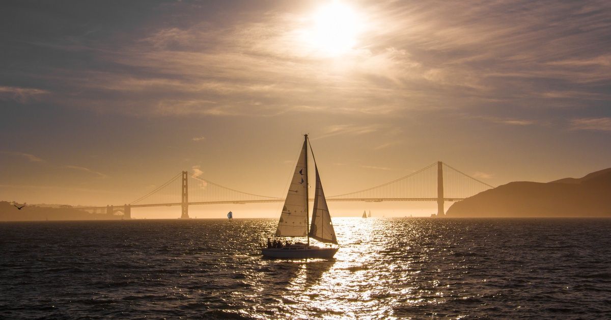 A sailboat is in the San Francisco Bay on a hazy day. The Golden Gate Bridge and additional boats are in the background.