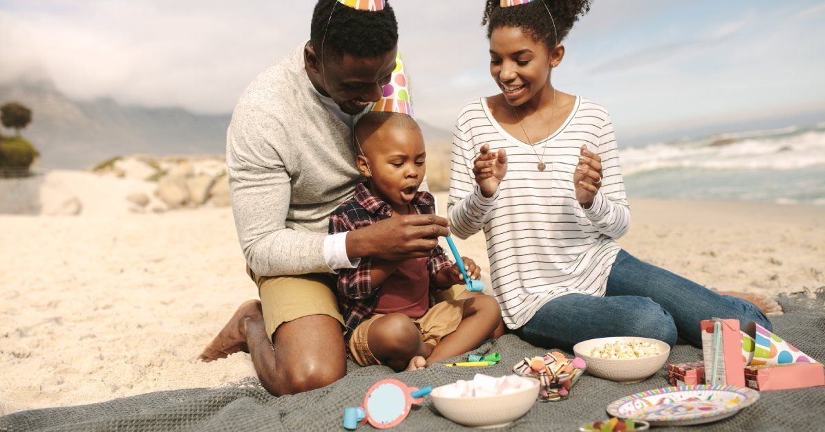Two adults and a child sit on the beach wearing party hats. In front of them are snacks, plates, and colorful noisemakers.