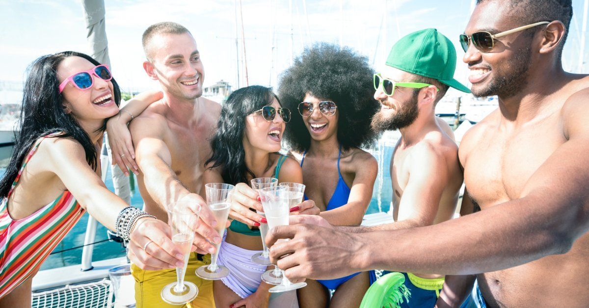 Smiling young men and women in swimsuits stand on a sailboat and bring their champagne glasses together for a toast.