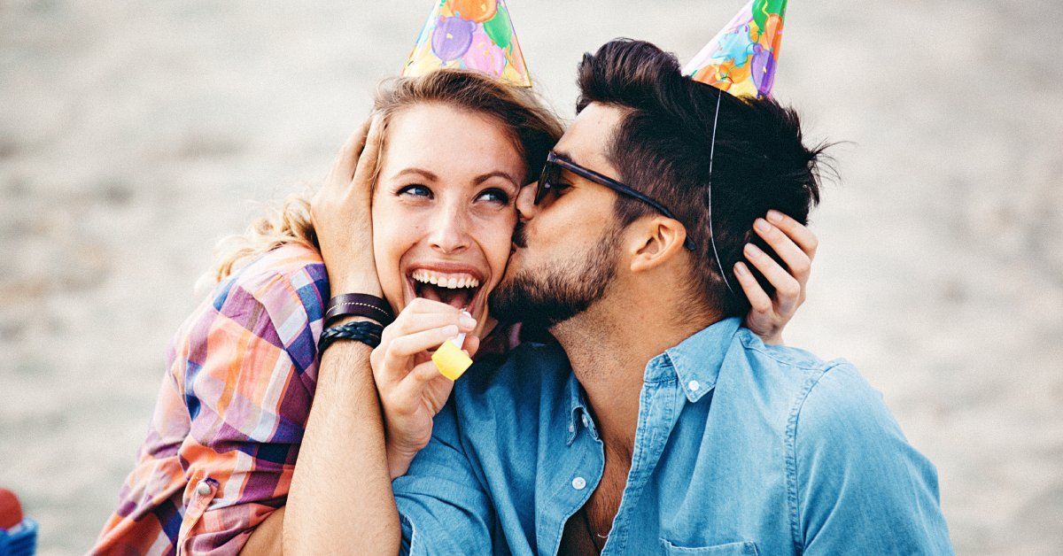 A young man and woman sit on the beach wearing party hats. He kisses her cheek while she smiles.