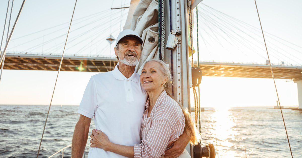 An older adult couple hugging on a sailboat as the sun sets behind them. They wear collared shirts and have graying hair.