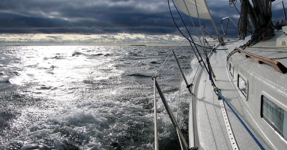 A white sailboat with no visible passengers sailing toward a gray, overcast sky and turbulent waves.